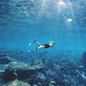 A person wearing flippers swims underwater over a large coral reef, with rays of sunlight filtering through the clear blue water.