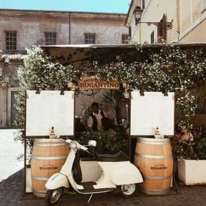 A white scooter is parked on a cobblestone street in front of an outdoor restaurant stand with two large wooden barrels.