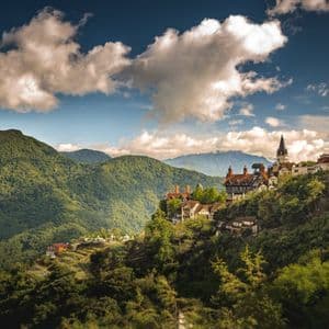 A European-style castle and village nestled in lush green mountains under a partly cloudy sky.