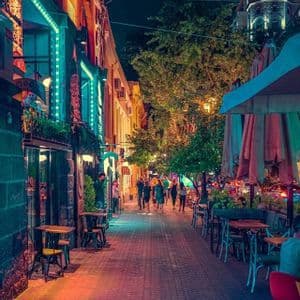 A narrow city street at night where people walk past outdoor cafes and under trees illuminated by colorful neon lights.