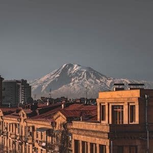 A large, snow-capped mountain rises in the distance behind the sunlit rooftops of a city under a hazy sky.
