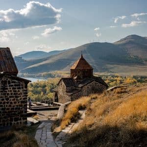 A winding stone path on a grassy hill leads to an old stone church, with mountains and a lake in the distance.