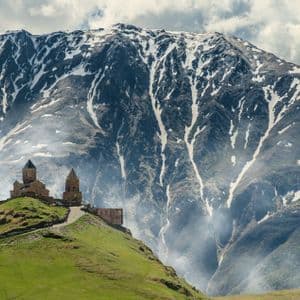 A stone church with towers on a grassy hill, with a large, snow-dusted mountain in the background.