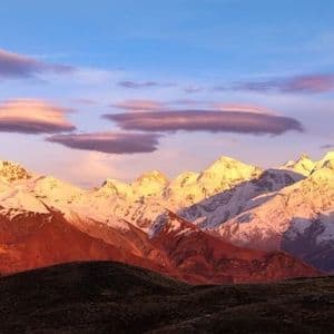 A snow-capped mountain range is illuminated by the golden light of sunrise against a blue sky with thin purple clouds.