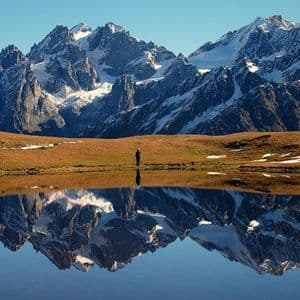 A person stands on a grassy shore before a still lake, which reflects a large, snow-covered mountain range under a clear sky.