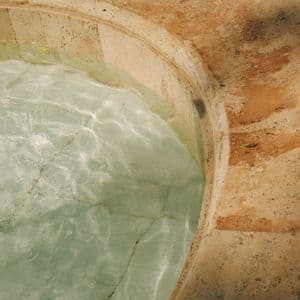 A close-up of a curved stone fountain edge with clear water inside, where sunlight creates reflections on the tiled bottom.