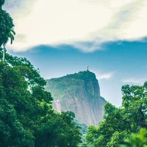 La estatua del Cristo Redentor se alza sobre una exuberante montaña verde, enmarcada por árboles en primer plano bajo un cielo nublado.