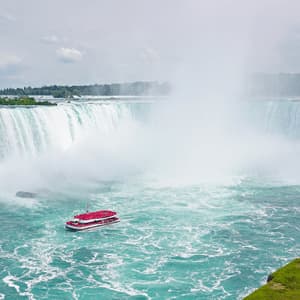 Una barca turistica rossa naviga le acque turchesi e turbolente ai piedi di una cascata imponente, che genera una fitta nuvola di nebbia.