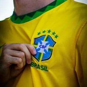 Primo piano della mano di una persona che tiene lo stemma della CBF sulla sua maglia da calcio gialla del Brasile.