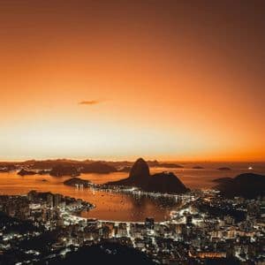 Una città costiera illuminata si curva attorno a una baia, accanto a un'imponente montagna conica, sotto un vibrante cielo arancione al tramonto.