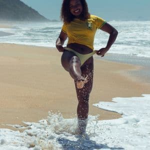 Una donna sorridente con una maglietta gialla del Brasile solleva acqua spumosa con il piede su una spiaggia soleggiata e sabbiosa.
