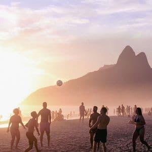 Sagome di un gruppo di persone che giocano a palla su una spiaggia sabbiosa al tramonto, con montagne lontane oltre l'acqua.