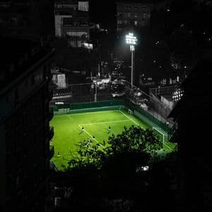 Veduta aerea di una partita di calcio su un campo verde brillantemente illuminato di notte, circondato da edifici scuri della città.