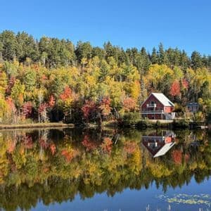 Una casa rossa e una fitta foresta con colori autunnali si riflettono perfettamente nell'acqua calma di un lago.