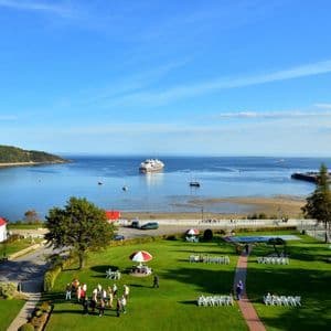 Vista panoramica di un borgo costiero con una chiesa dal tetto rosso, un prato verde e una grande baia con una nave da crociera.