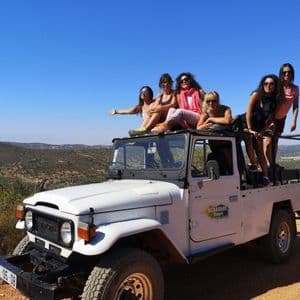 Un viaje en grupo de WeRoad de mujeres posando en un jeep de safari blanco en un camino de tierra, con un paisaje montañoso bajo un cielo azul claro.
