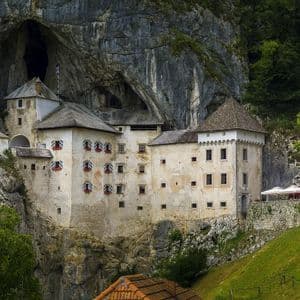 Un grande castello bianco, incastonato direttamente all'imbocco di una grotta buia su una parete rocciosa a picco, circondato da una lussureggiante vegetazione verde.