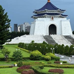 Ein weißes, traditionelles Gebäude mit einem blauen Stufendach liegt hinter einem üppigen, angelegten Garten mit gepflegten Hecken und grünen Rasenflächen.