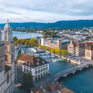 An aerial view of a historic city with twin cathedral towers, a river with a bridge, and boats docked along the waterfront.