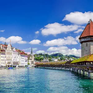A wooden covered bridge with a stone tower crossing a river next to a historic city waterfront under a blue sky.