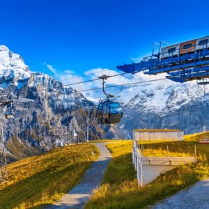 A cable car travels over a green mountain slope, with towering snow-capped peaks visible against a clear blue sky.