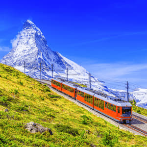 An orange train travels along a track on a green mountainside, with a large, snow-covered mountain peak in the background.