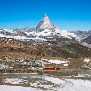 A red train travels on a track across a rocky, partially snow-covered mountainside, with a prominent snow-capped peak in the background.
