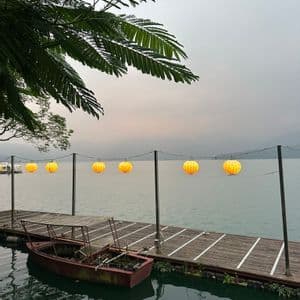 Leuchtende gelbe Papierlaternen hängen über einem Holzsteg, an dem ein kleines Boot auf einem ruhigen See vertäut ist.