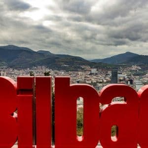 Un grande cartello rosso con la scritta 'Bilbao' da un punto panoramico che domina la città, con le montagne sullo sfondo e un cielo nuvoloso.