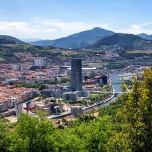 Una veduta panoramica di una grande città con un fiume serpeggiante, incastonata tra dolci colline verdi e montagne sotto un cielo blu.
