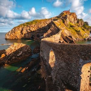 Un ponte di pietra tortuoso con archi attraversa l'acqua fino a un isolotto roccioso con un edificio sulla cima, sotto un cielo azzurro nuvoloso.