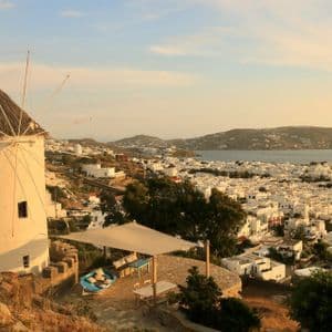 Un mulino a vento bianco tradizionale su una collina che domina una cittadina costiera con edifici bianchi e il mare durante un tramonto dorato.