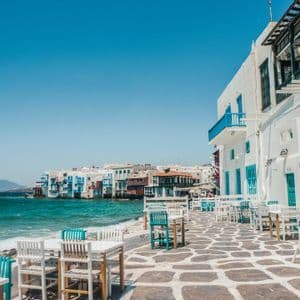 Edifici imbiancati a calce con dettagli blu costeggiano un patio in pietra con tavoli da caffè all'aperto accanto al mare turchese.