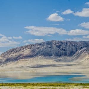 Un lac turquoise paisible s'étend au pied d'une vaste montagne aride, sous un ciel bleu partiellement nuageux.