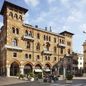 Ein verziertes Backsteingebäude mit Bogenfenstern und einem Eckerker auf einem gepflasterten Stadtplatz unter blauem Himmel.