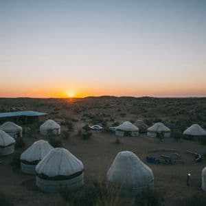 Un camp de yourtes traditionnelles s'étend sur un paysage désertique au coucher du soleil, avec le soleil rougeoyant à l'horizon.