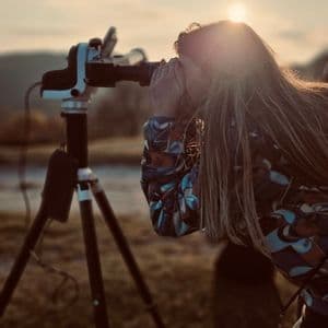 Una persona con i capelli lunghi guarda attraverso una fotocamera montata su un treppiede, all'aperto al tramonto, con il sole che crea un bagliore.