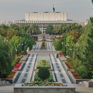 Une large allée symétrique de parc, bordée d'arbres et de parterres de fleurs colorés, mène vers un grand bâtiment moderne et blanc au loin.