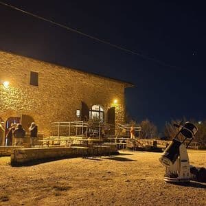 Un grande telescopio si erge in una radura ghiaiosa di notte, puntato verso il cielo, con un gruppo WeRoad riunito fuori da un edificio in pietra illuminato.