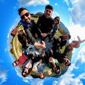 Un viaggio di gruppo WeRoad che sorride per un selfie a 360 gradi effetto 'piccolo pianeta' in un paesaggio verde e collinare sotto un cielo azzurro.