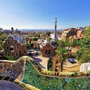 Un colorato muro a mosaico in primo piano domina un parco con architettura unica e un lontano skyline cittadino sotto un cielo blu.
