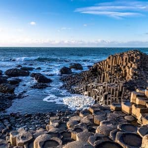 Colonne esagonali di basalto incastrate formano un paesaggio costiero, con le onde dell'oceano che lambiscono la riva sotto un cielo azzurro.