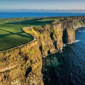 Una vista aerea di scogliere a picco che si gettano nell'oceano blu profondo, accanto a vasti campi verdi sotto un cielo soleggiato.