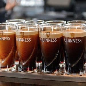 Un primo piano di diversi boccali di birra Guinness appena versati, con cremose schiume, disposti in fila su un vassoio metallico da bar.