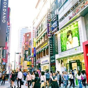 Una folla di persone cammina lungo una trafficata strada cittadina affiancata da alti edifici con numerose insegne colorate e vetrine di negozi.