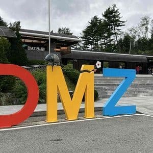 Grandi lettere colorate che compongono la scritta DMZ si ergono di fronte a un padiglione espositivo, con un fiore bianco che spunta dalla 'M' spezzata.