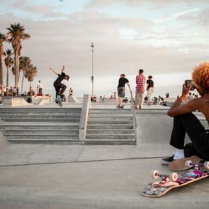 Una persona riprende con il telefono mentre un altro skater esegue un trick a mezz'aria in uno skatepark di cemento con palme.