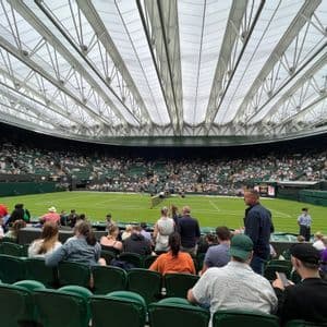 Una grande folla siede sui seggiolini verdi dello stadio guardando una partita di tennis su un campo in erba sotto un grande tetto retrattile.