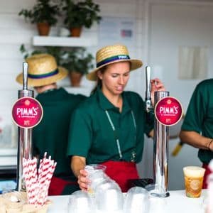 Bartender in uniformi verdi e cappelli di paglia versano bevande dai rubinetti di Pimm's & Lemonade a una bancarella.