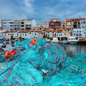 Un mucchio di reti da pesca blu intenso con galleggianti colorati si trova su un molo di fronte a un porto con barche ed edifici sul lungomare.